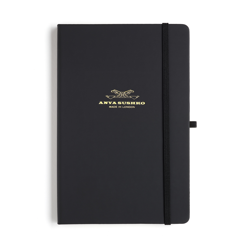 A black hardback A5 refill Luxury notepad with a gold embossed brand logo on the cover, a ribbon bookmark, and an elastic pen holder.