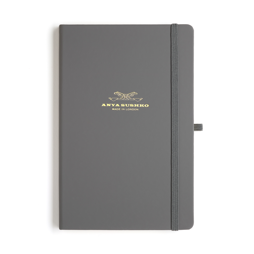 A gray A5 hardback refill Luxury notepad with a gold embossed brand logo at the front and a ribbon bookmark.