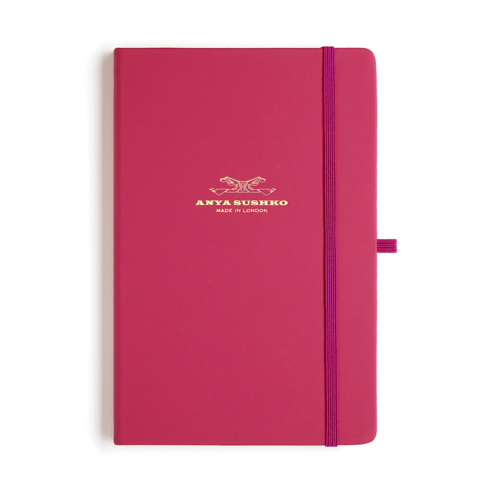 A pink A5 hardback refill Luxury notepad with a gold embossed brand logo at the front and a purple elastic pen holder.